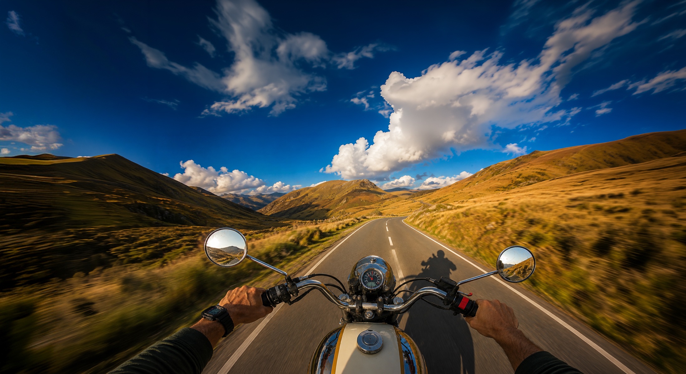 First-person motorcycle view riding through a mountain road at sunset - handlebars, mirrors, and speedometer with the road stretching into the hills.